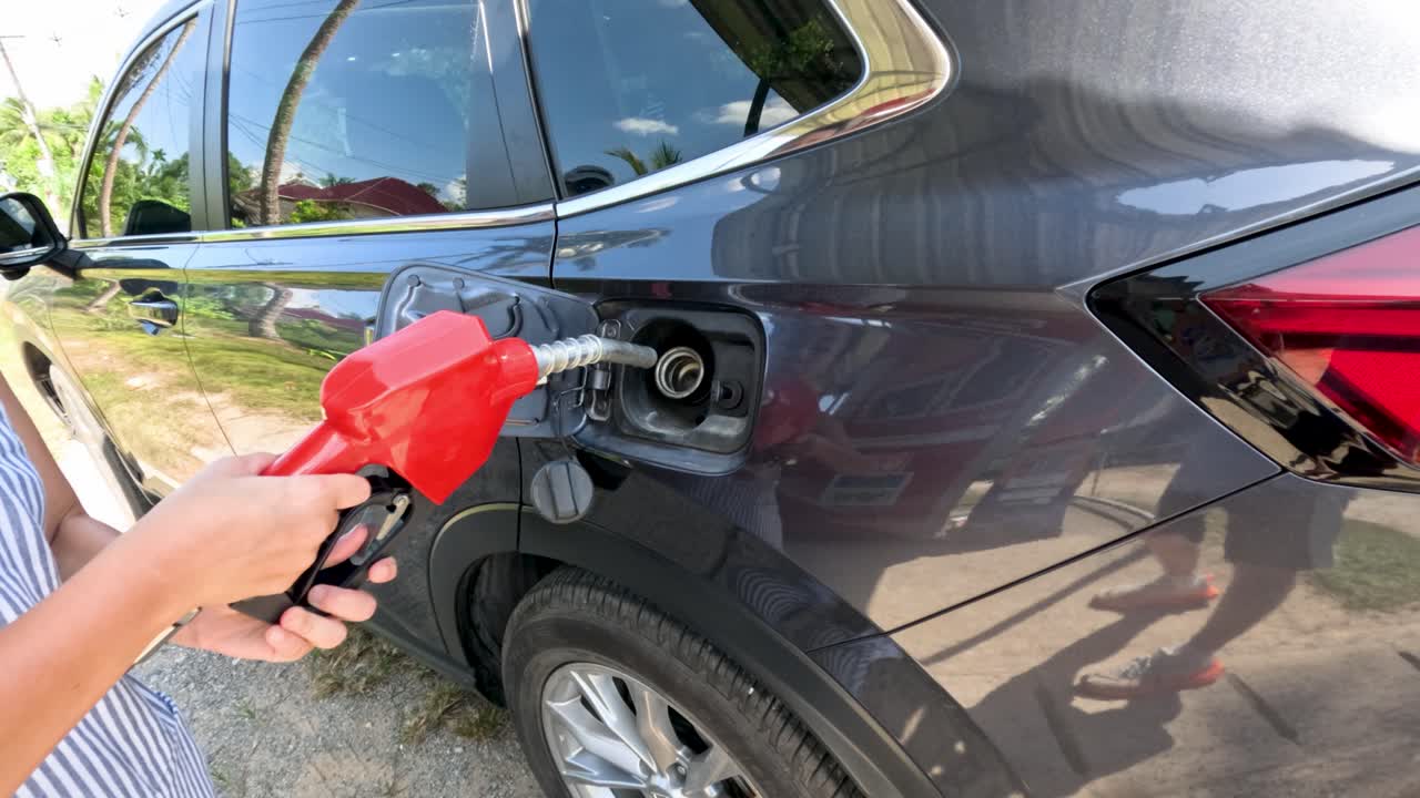 Person inserts red fuel nozzle into black SUV under bright daylight, static camera, outdoor setting