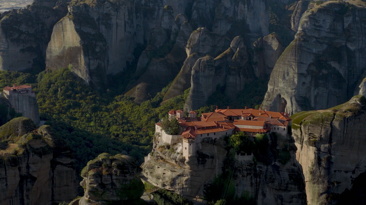 Vista aerea cinematografica del monastero di Meteora in Grecia, arroccato su imponenti scogliere, formazioni rocciose suggestive e una lussureggiante valle verde creano una scena storica mozzafiato