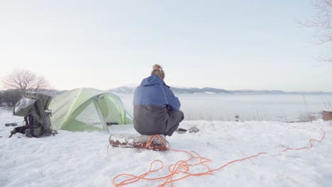 guy sitting on a log next to the campfire outside his tent at winter