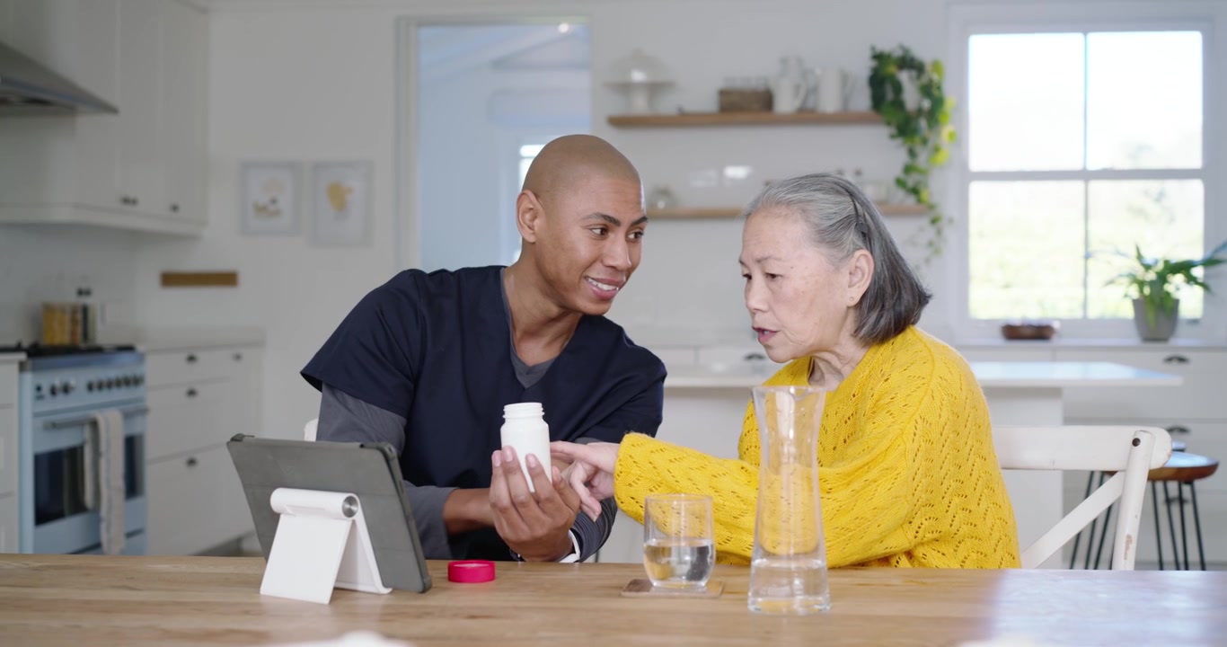 Caregiver in scrubs showing pill-label on tablet to Senior Chinese woman at kitchen explaining meds