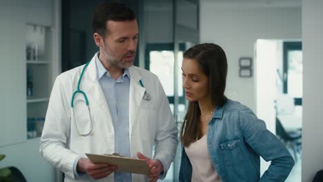un médico caucásico y una paciente discuten documentos en el pasillo de un centro de salud.