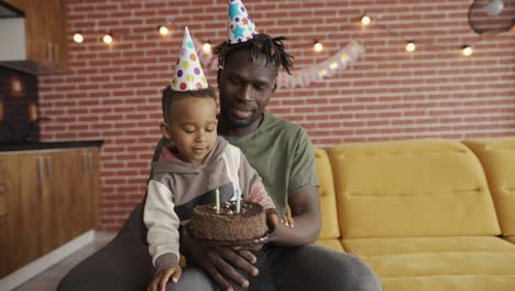 padre afroamericano tiene in braccio il figlio - soffia tre candele sulla torta di compleanno
