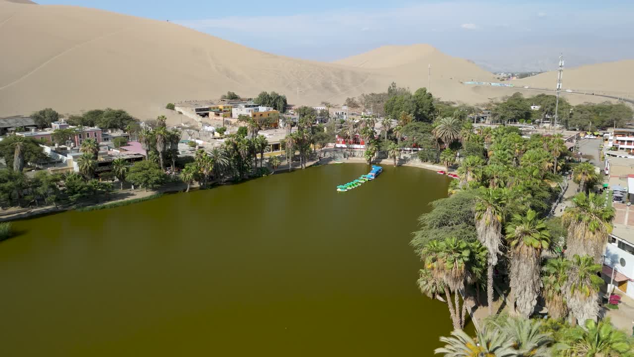 Aerial parallax shot of the Huacachina desert oasis with lake and buildings in Ica, Peru, surrounded by palm trees and large sand dunes
