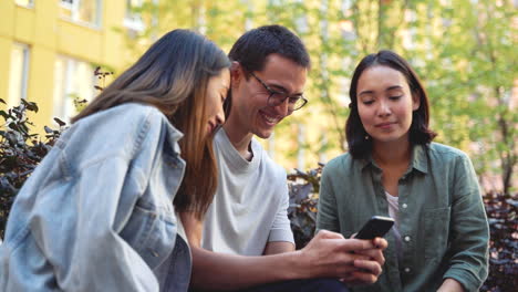 group of three young japanese friends looking something funny on mobile phone and laughing while sitting together in the park