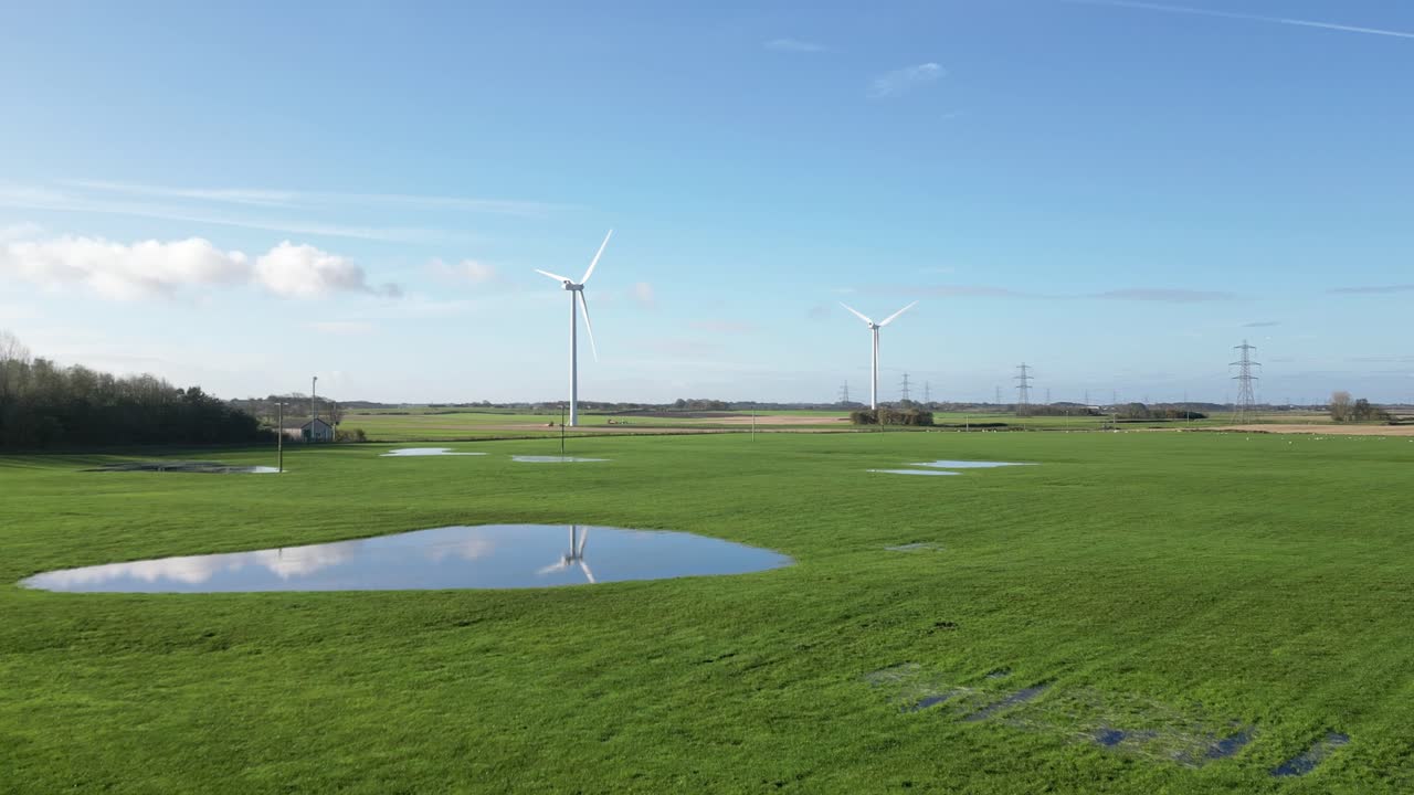 Slow-moving drone shot across a farm field, heading toward two wind turbines with reflection in a puddle. The rural landscape and blue skies create a stunning backdrop for sustainable energy