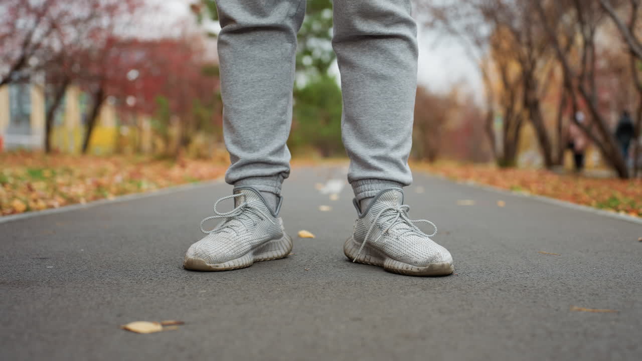 Leg view of sports man in jogger and sneakers standing still as wind blows dry foliage around on park path, surrounded by autumn colors and fallen leaves