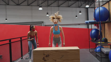 caucasian woman jumping on box wearing face mask at gym, mixed race woman watching