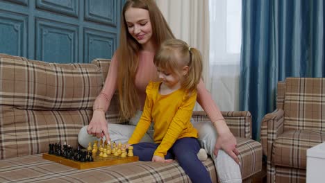 Small-cute-daughter-child-and-young-mother-playing-chess-at-home-sofa,-leisure-hobbies-activities