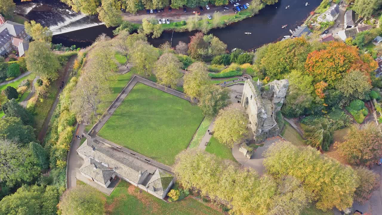 High angle top down drone view of rectangular lawns, surviving stone walls and ruins of Knaresborough Castle, surrounded by mature autumn trees and overlooking the River Nidd weir in the city centre