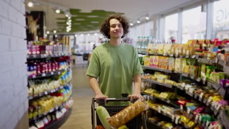 un chico moreno con cabello rizado camina con un carro lleno de comestibles y mira las mercancías en un supermercado de comestiables