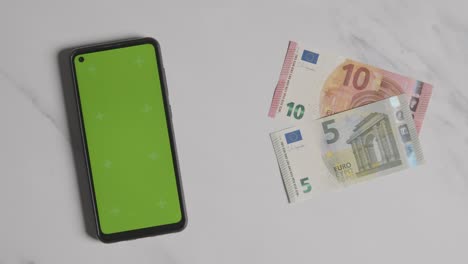 Overhead-Currency-Shot-Of-Hand-Grabbing-10-And-5-Euro-Notes-Next-To-Green-Screen-Mobile-Phone