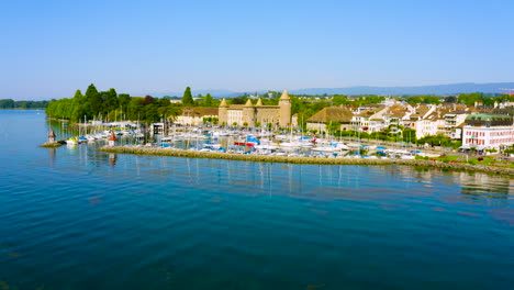 Boats-Anchored-On-The-Harbor-By-The-Lake-Leman-With-Morges-Town-And-Castle-On-The-Lakeshore-In-The-Canton-Of-Vaud-In-Switzerland---low-aerial-drone