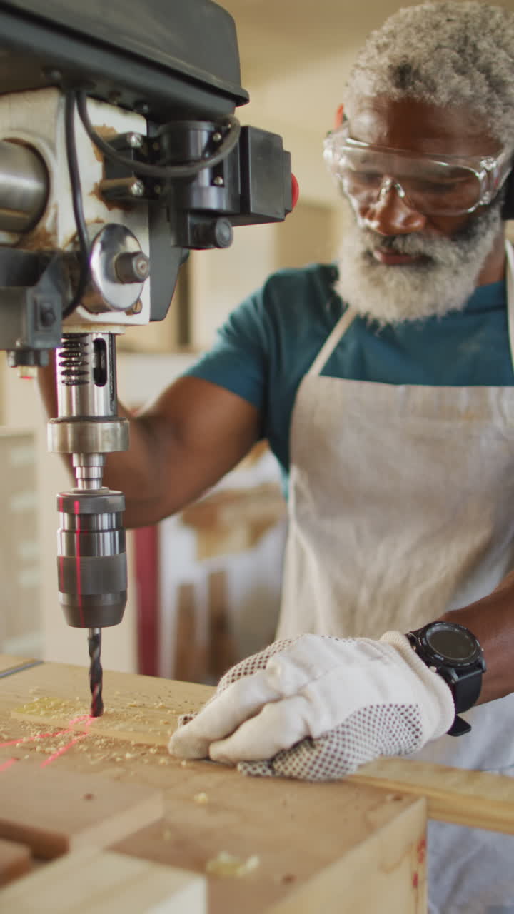 Vertical video of senior african american male carpenter drilling in workshop