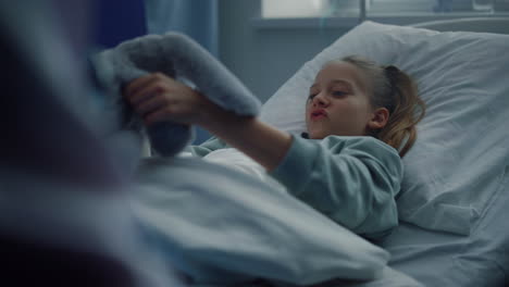 sick girl playing soft toy in clinic bed. kid lying in modern hospital room.