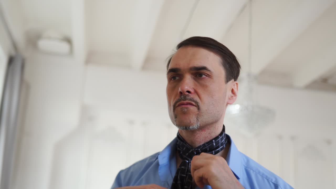 un hombre con una camisa azul ata una corbata con sus manos cerca de un espejo en una gran habitación blanca