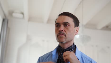 un hombre con una camisa azul ata una corbata con sus manos cerca de un espejo en una gran habitación blanca