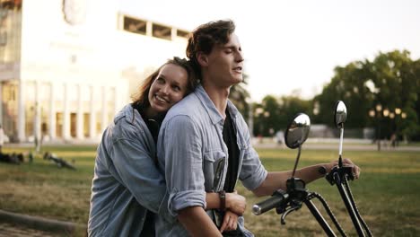 Young-attractive-couple-hugging-and-enjoying-themselves-while-sitting-on-a-bike-on-the-street.-Both-wearing-blue-shirts.-Sunlight