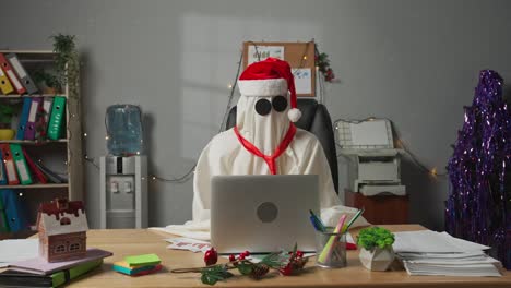 a 'ghost' figure wearing a santa hat works on a laptop in a christmas-decorated office