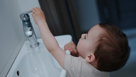 little caucasian baby reaches hand to push the tap. happy kid touches water smiling excitedly. close up.