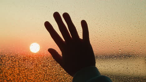 hand silhouette on window covered with raindrops closeup palm touches wet glass against bright red