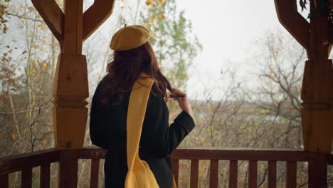 back view of a lady in a yellow beret playfully twisting her hair while standing close to a wooden railing, gazing out into the autumn woods