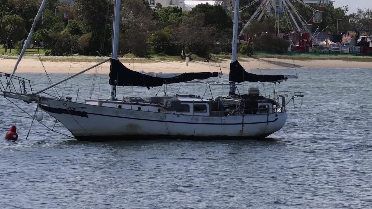 A sailboat rests calmly on the water near a sandy beach with trees in the background.