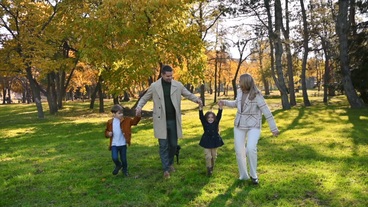 Happy family in an autumn park. Mother and father slowly running with daughter, son and their dog, yellowed trees around. Slow motion