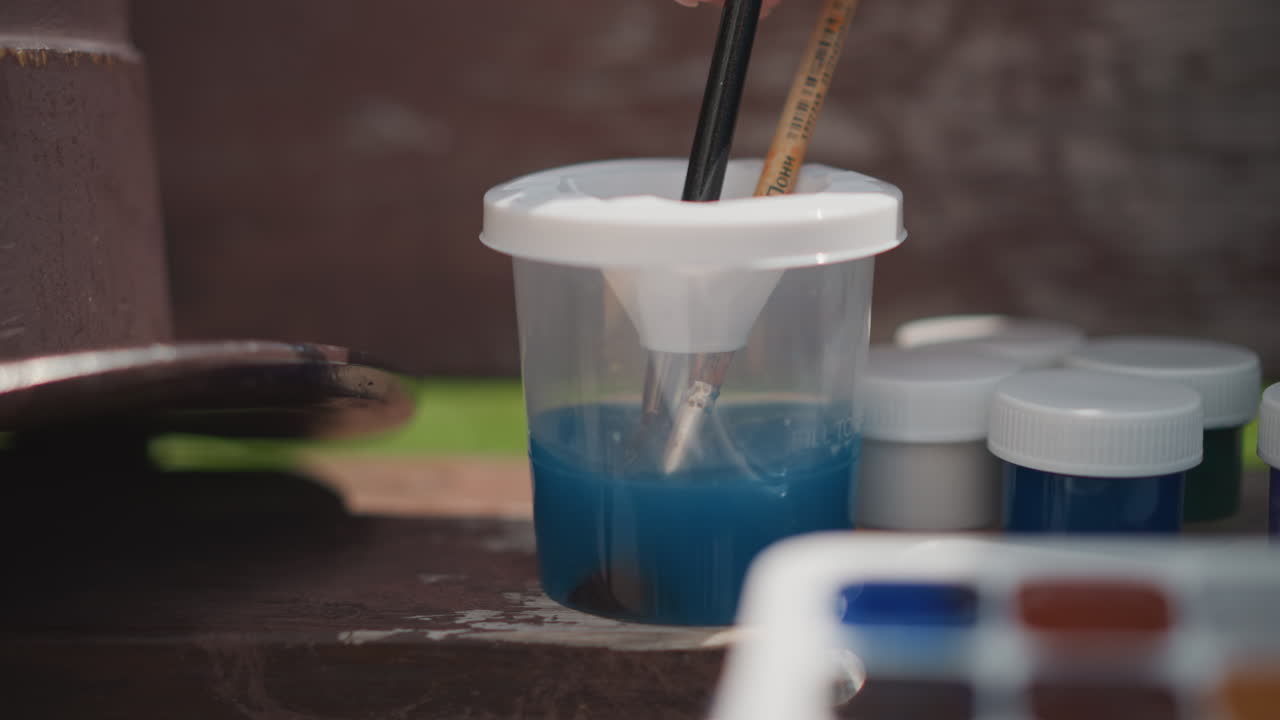 Cat biru dalam cangkir dengan kuas. Pemandangan meja luar ruangan ini menampilkan cangkir pencampur plastik berisi cat air biru tua, dengan kuas-kuas terendam. Toples cat dan palet di dekatnya terlihat buram, diterangi cahaya alami yang lembut dengan kedalaman fokus dangkal.