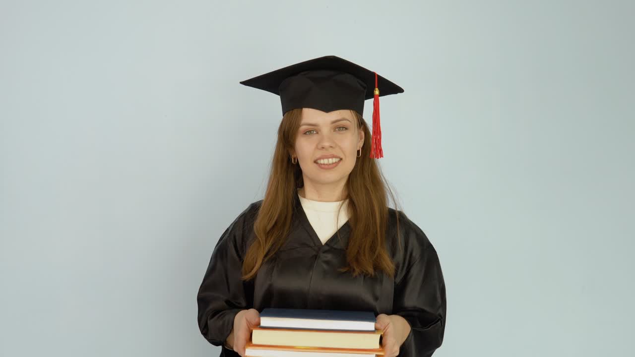 una joven mujer caucásica con una túnica negra y un sombrero de maestro se mantiene erguida sosteniendo libros de texto y mira a la cámara. retrato de media altura