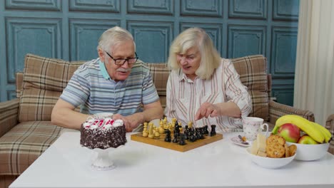 Retired-senior-couple-talking-drinking-tea,-playing-chess-in-modern-living-home-room-lounge-together