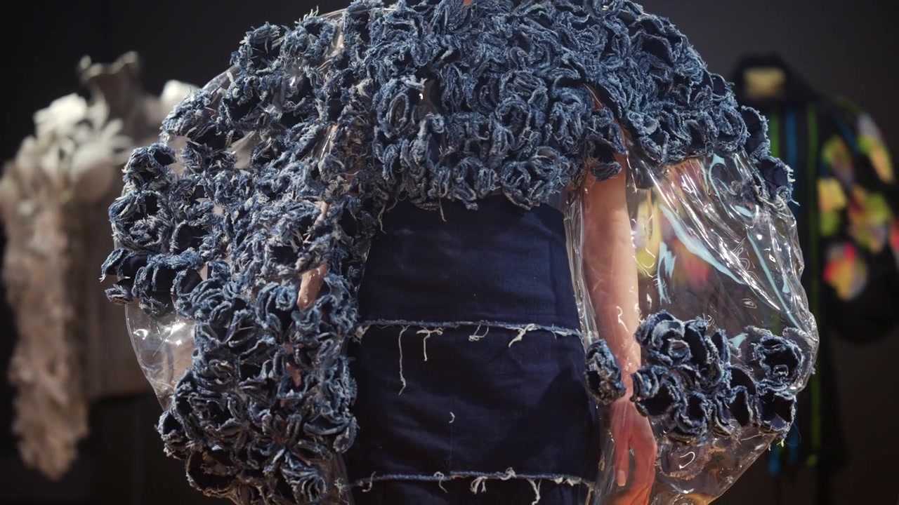 View of a model wearing an avant garde dress featuring textured blue fabric elements and transparent oversized sleeves