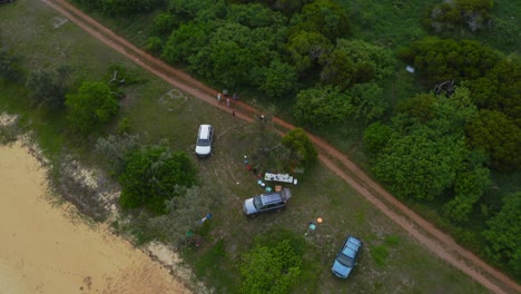 Un-Dron-Captura-Una-Foto-De-Personas-Disfrutando-De-Un-Picnic-Cerca-De-Un-Camino-De-Tierra,-Mostrando-El-Paisaje-Natural-Que-Los-Rodea,-Incluida-La-Playa-De-Arena-Y-La-Exuberante-Vegetación-De-Los-árboles-Durante-El-Día