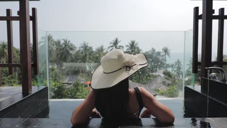 Back-view-of-a-female-tourist-wearing-a-sun-hat-and-sitting-in-a-open-air-bath-on-a-balcony-or-terrace-overlooking-a-tropical-island-of-Koh-Kood-in-Thailand