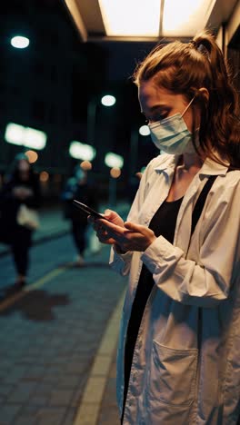 young woman standing at nighttime bus stop, wearing protective face mask while using smartphone, illuminated by streetlights with calm, pensive demeanor during quiet evening commute