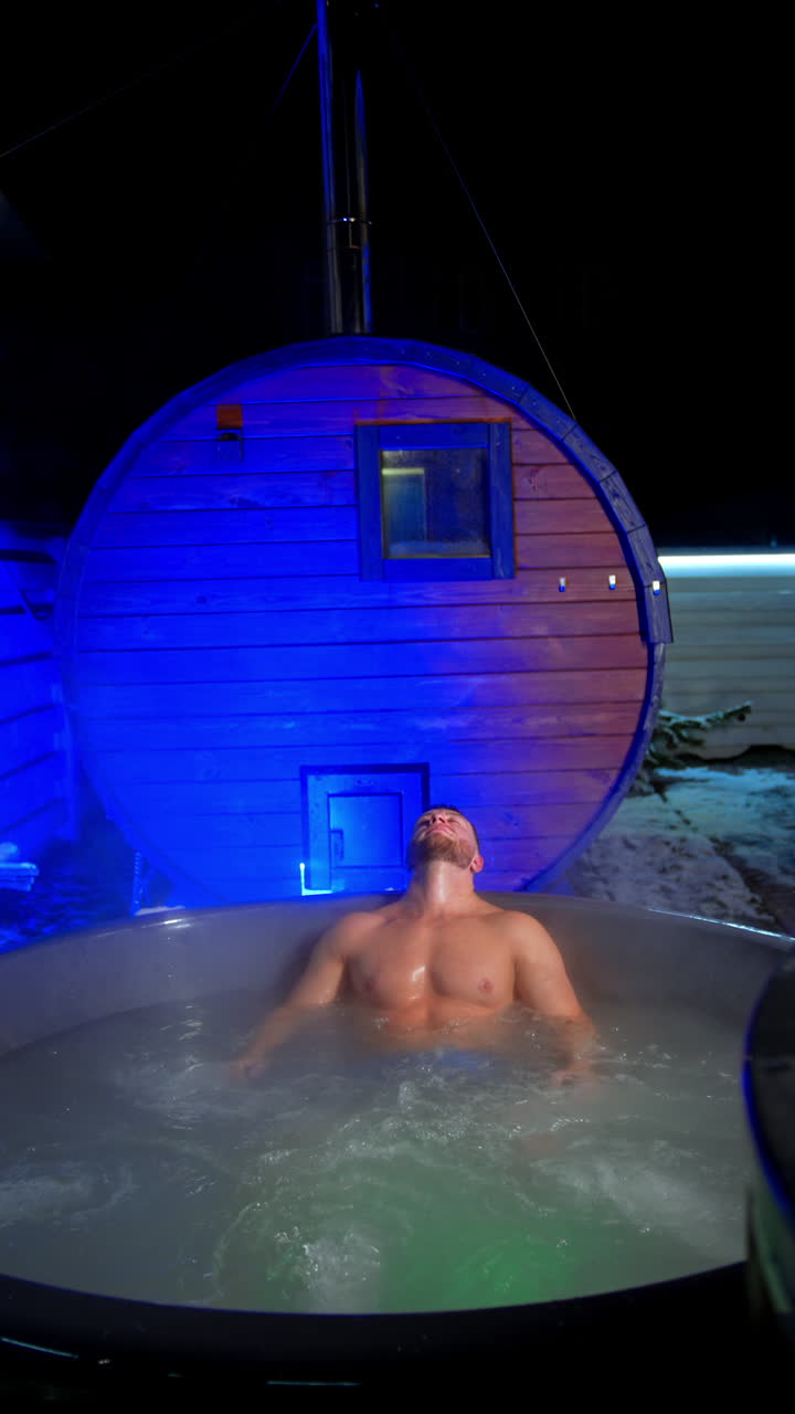 Strong muscular Caucasian man sits in hot tub holding his hands to the sides of the tub. Relaxation time in spa outdoors in winter. Vertical video.