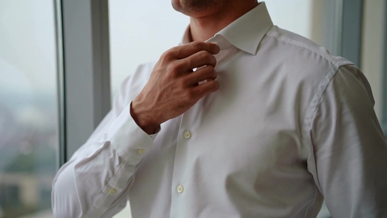 Uomo che si sistema, con camicia bianca elegante, che liscia il colletto e la parte anteriore vicino a una finestra alta, preparandosi per una riunione.