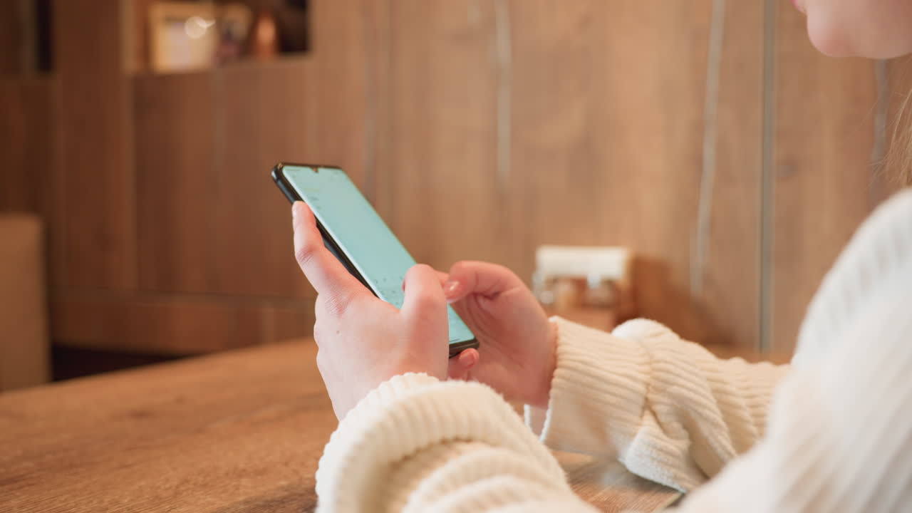Close up hand view of white woman in ribbed sweater operating smartphone on wooden cafe table, focused on screen while seated in warm indoor setting with blurred background