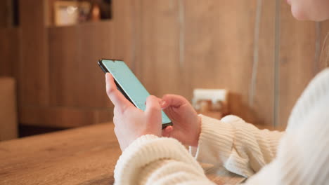 close up hand view of white woman in ribbed sweater operating smartphone on wooden cafe table, focused on screen while seated in warm indoor setting with blurred background