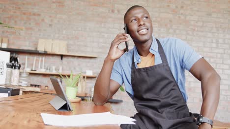 happy african american male coffee shop owner using tablet talking on smartphone, slow motion
