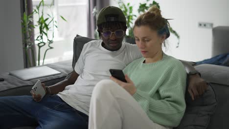 happy biracial couple relaxing on sofa with smartphone