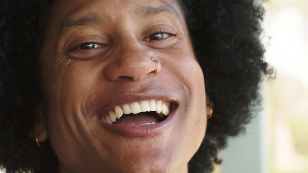 Man in 30s wearing gold hoops and nose ring shifting to laughter by balcony window