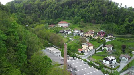 drone going up in front of small factory chimney with settlement between green nature in back