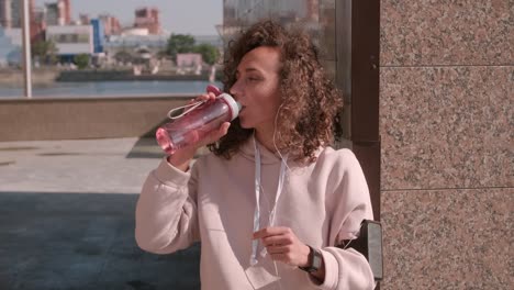 chica bastante rizada bebiendo agua después de hacer ejercicio al aire libre