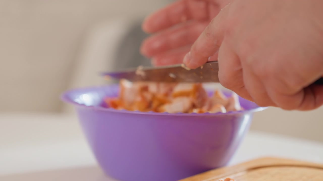 Hands gently press, lift, and drop diced filling into purple plastic bowl on wooden cutting board in warm indoor kitchen, showing careful food preparation, mixing, and assembly for homemade meal