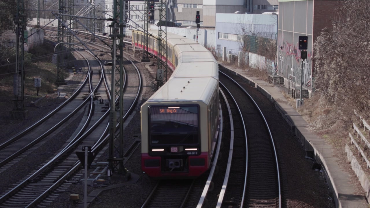 Una vista dall'alto mostra un treno S-Bahn che viaggia lungo i binari ferroviari in un contesto urbano. Pali in acciaio per la linea aerea di contatto si ergono in primo piano mentre il treno curva lungo i binari in una giornata limpida.