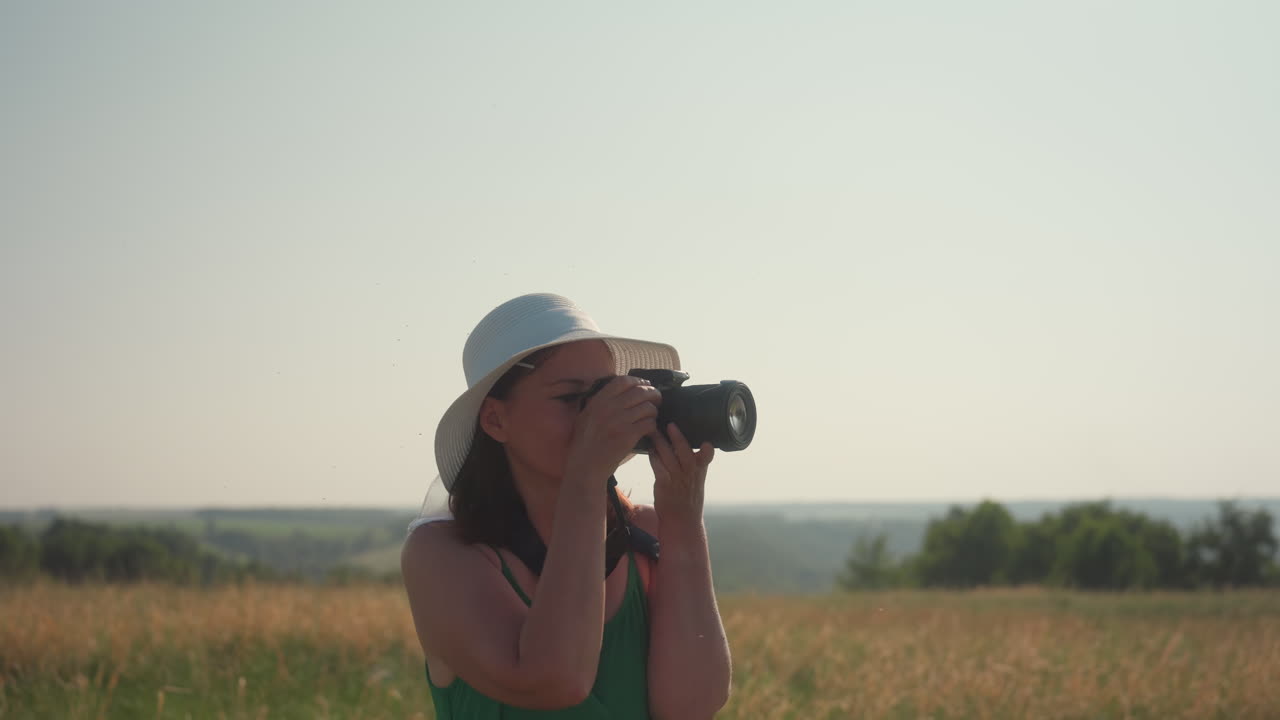 Woman in white sunhat and green dress with camera walks through tall countryside grass, admiring landscape under warm daylight, enjoying peaceful nature, photography