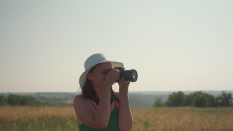 woman in white sunhat and green dress with camera walks through tall countryside grass, admiring landscape under warm daylight, enjoying peaceful nature, photography