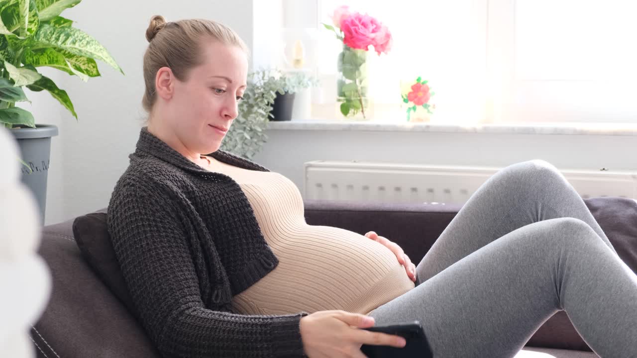 Pregnant white woman sitting on the couch typing on phone while smiling and caressing her belly by daylight slow motion