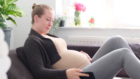 pregnant white woman sitting on the couch typing on phone while smiling and caressing her belly by daylight slow motion