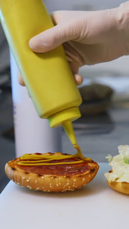 a hand in white gloves pouring sauce on toasted half of the bun. making a delicious hamburger. kitchen in restaurant. front video. vertical video